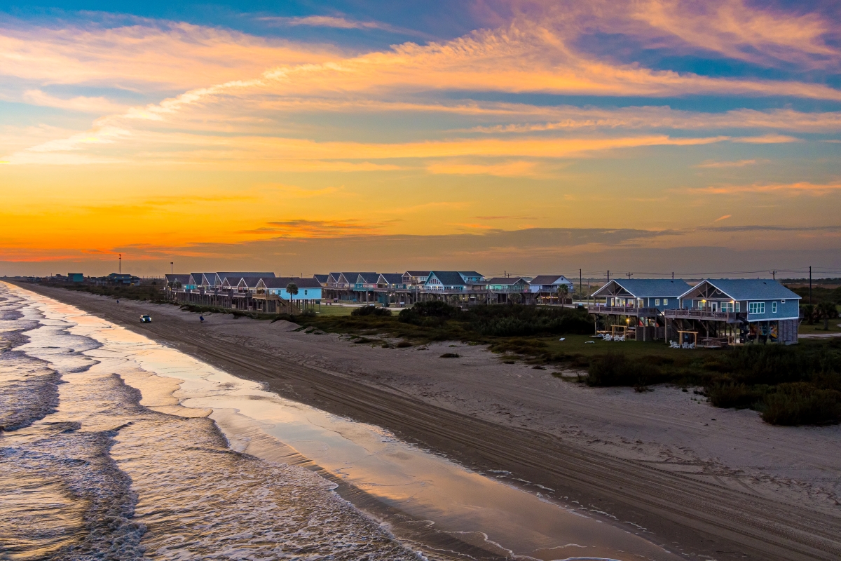 Aerial view of a row of stilted beach houses along a drive-on sand beach at sunset with waves rolling in