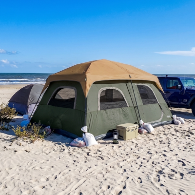 A large tent staked out on white sand next to a Jeep, with sandbags anchoring the corners and a cooler and lantern set up outside
