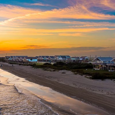 Aerial view of a row of stilted beach houses along a drive-on sand beach at sunset with waves rolling in