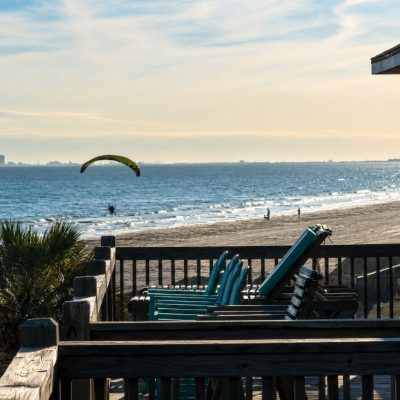 Beach house deck with stacked teal chairs facing a calm beach where a paraglider flies low over the water