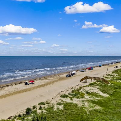 Aerial view of Bolivar Peninsula Texas showing trucks parked on the sand, beach access boardwalk, coastal dunes, and vacation homes along the shoreline